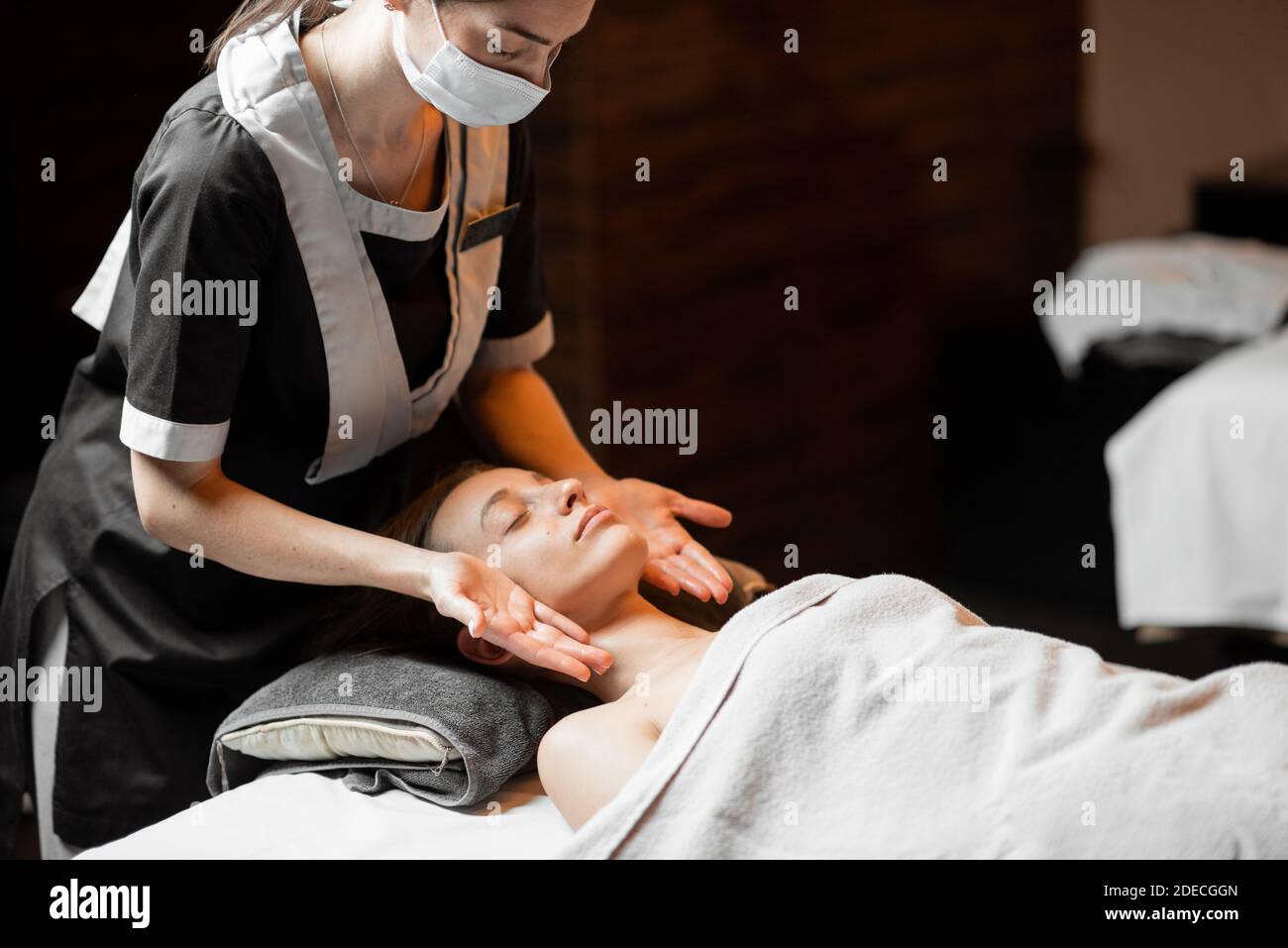 Massaggiatrice femminile professionista in maschera medica che fa massaggio facciale ad un cliente al salone di Spa. Business durante il concetto epidemico Foto Stock