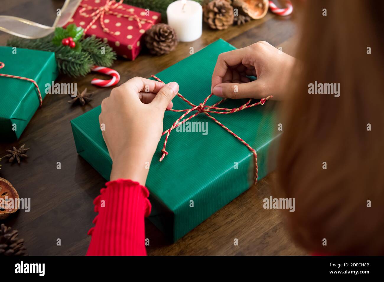 Donna che lega la stringa intorno alla scatola del regalo in carta di imballaggio di Natale verde come presente con gli articoli di decorazione sulla tabella di legno, che si prepara per celebrare festi Foto Stock