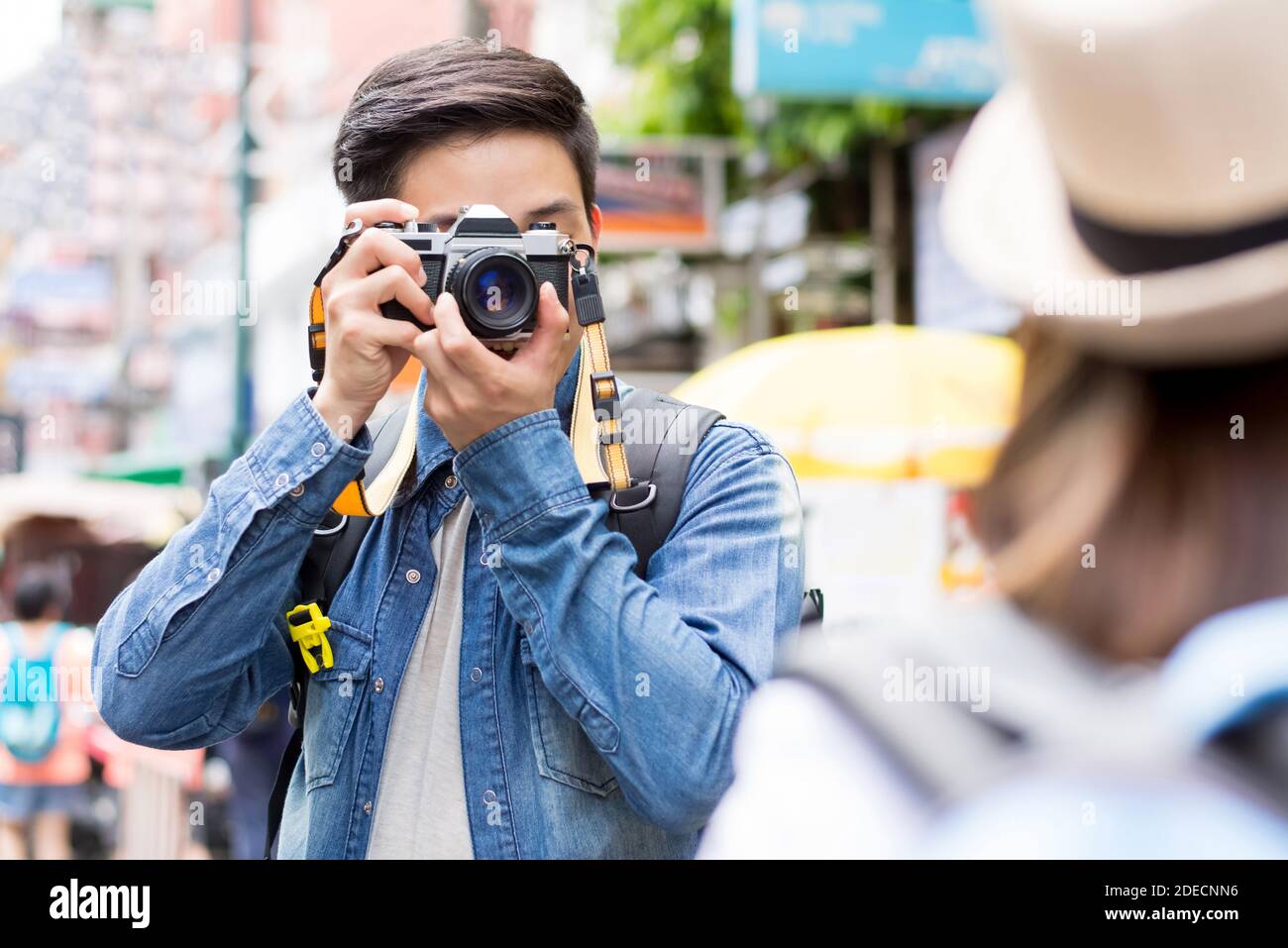 Fotografo turistico asiatico maschile in giacca blu denim scattando foto di compagno di viaggio femminile in posa in fuori città mercato di strada mentre in vacanza vis Foto Stock