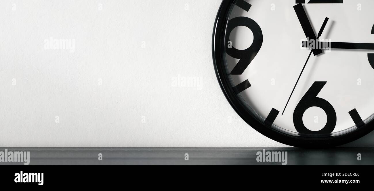 Orologio moderno minimalista o semplice bianco e nero su tavolo di legno nero e parete bianca con spazio per il testo. Banner orizzontale lungo Foto Stock