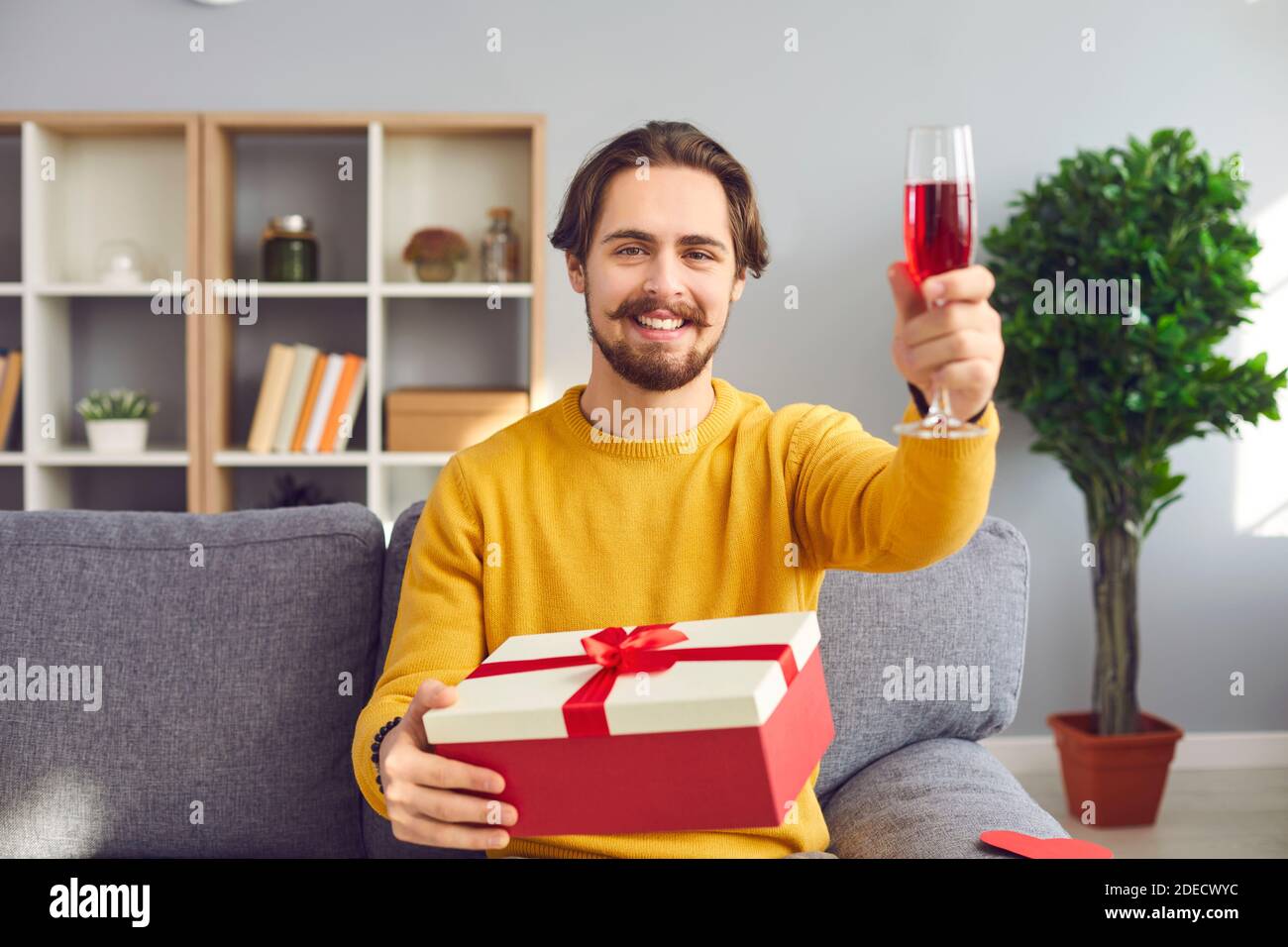 L'uomo hipster si siede con un regalo e solleva un bicchiere di vino rosso per un compleanno o San Valentino. Foto Stock