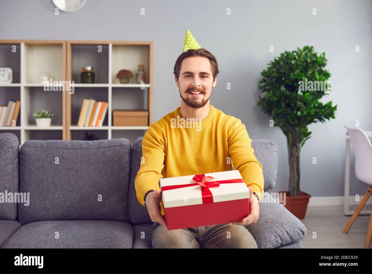 Giovane uomo positivo nel cappellino festivo seduto sul divano e mostrando la casella regalo per le festività Foto Stock