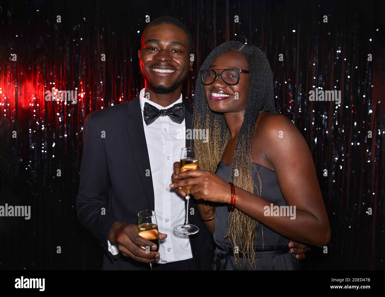 Ritratto in vita di una giovane coppia afroamericana che tiene bicchieri da champagne e sorride alla fotocamera mentre si posa su sfondo scintillante durante la festa, Foto Stock