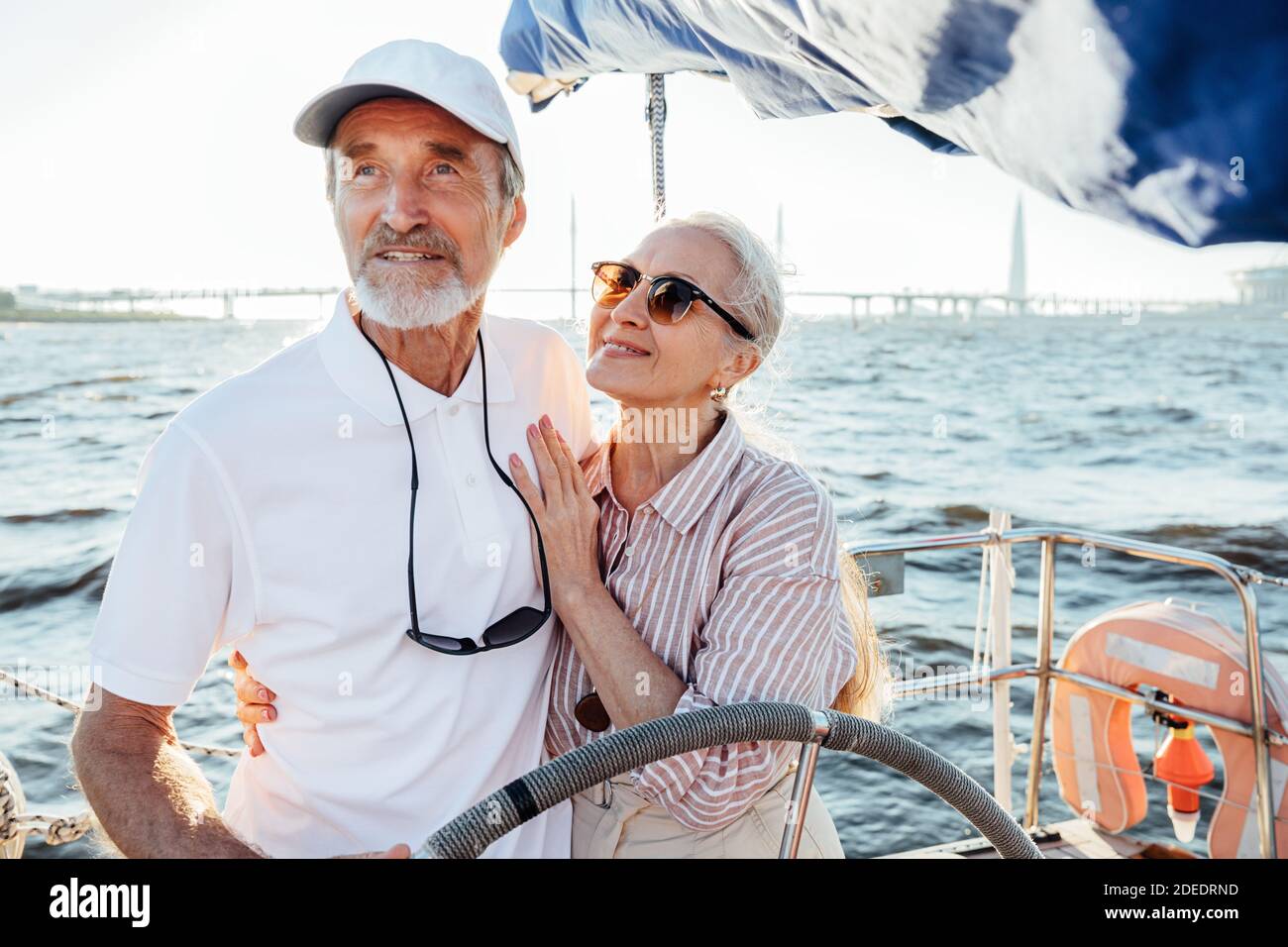 Donna matura in occhiali da sole abbracciando il marito e guardando lui mentre guida la barca a vela Foto Stock