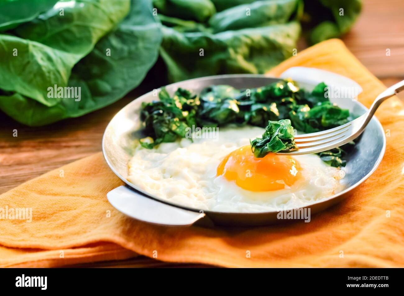 Spinaci verdi cotti con uova fritte su piastra in padella di ferro. Uova fritte sana prima colazione mangiare su sfondo tavolo di legno servito da tovagliolo arancione, forchetta Foto Stock