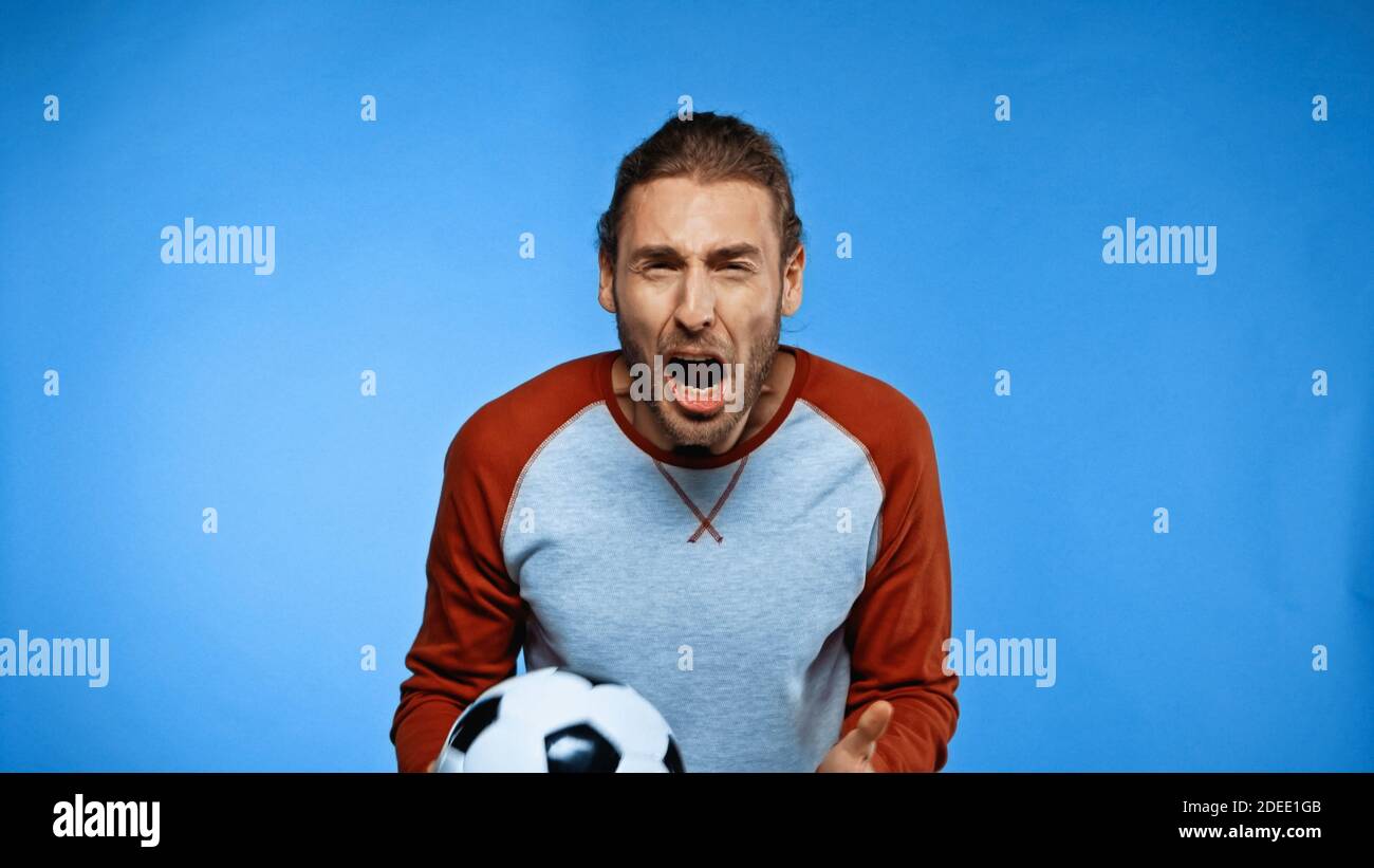 infuriato giovane tifoso di calcio che tiene la palla di calcio e urla su blu Foto Stock