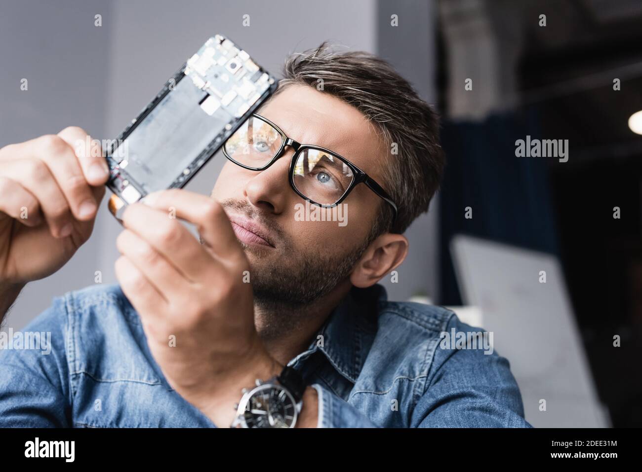 Addetto alla riparazione mirato negli occhiali che guarda il componente disassemblato del mobile telefono su sfondo sfocato Foto Stock