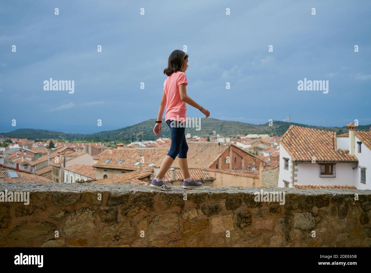 Giovane ragazza che indossa una blusa rosa che cammina su un muro di pietra con case sullo sfondo a Mora de Rubielos, Teruel, Spagna Foto Stock