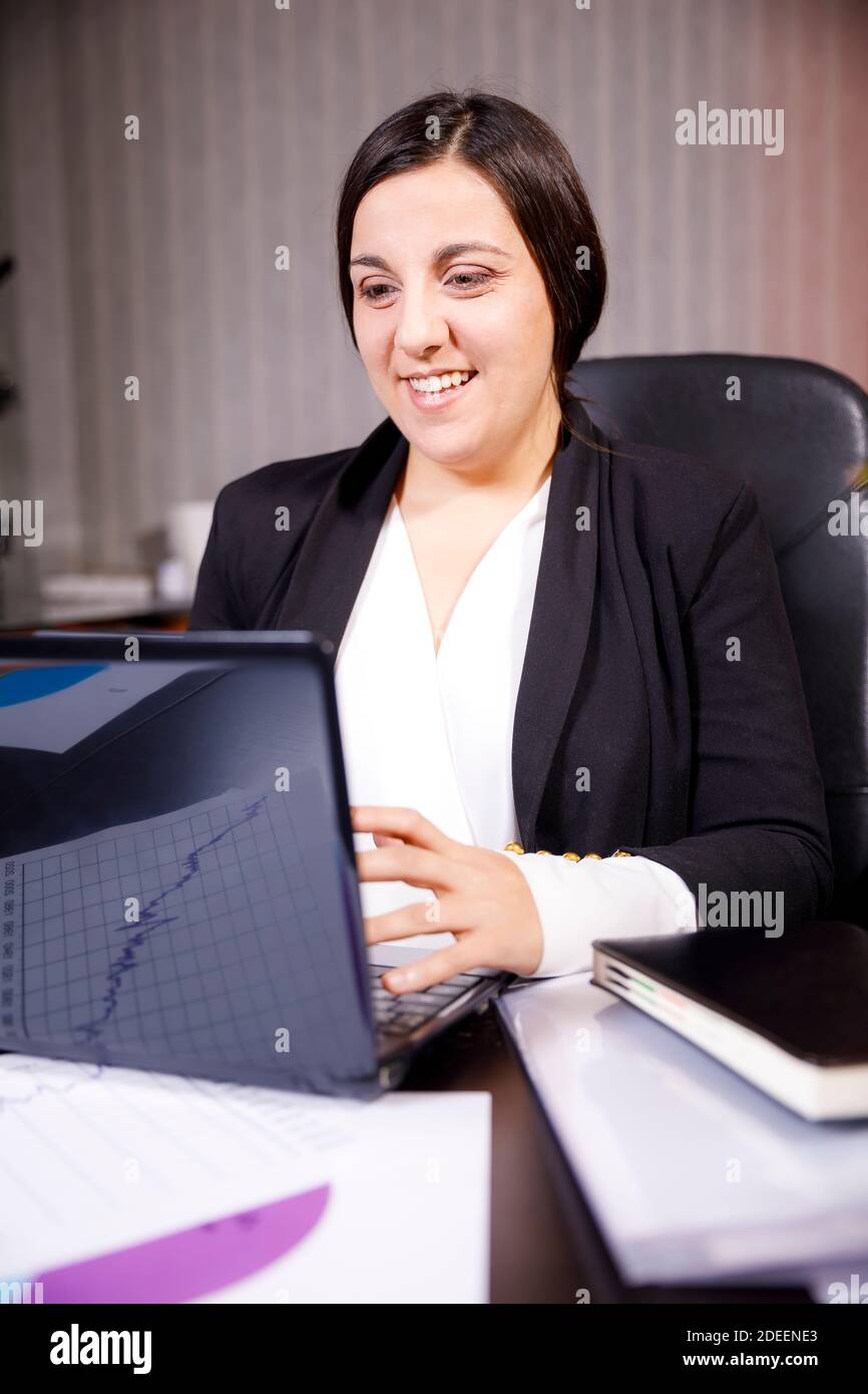 Giovane lavoratore di ufficio ragazza vestita in un vestito seduto a. desktop sorridente e utilizzando un computer portatile Foto Stock