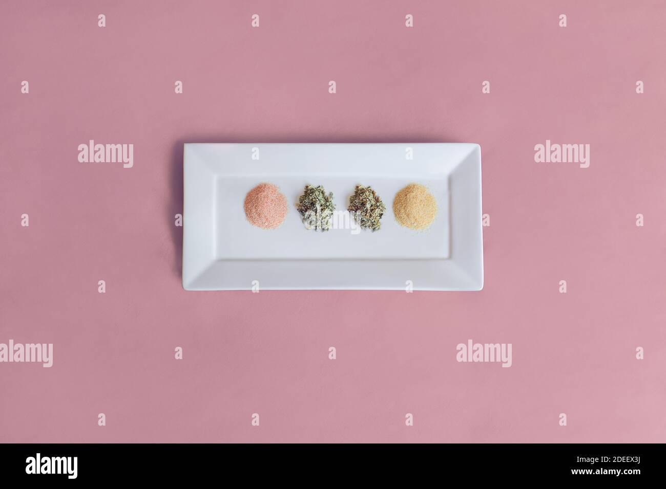 Un elegante piatto bianco posto al centro di un tavolo con una tovaglia rosa pastello contiene foglie secche e spezie in polvere per cucinare. Cibo sano. Foto Stock