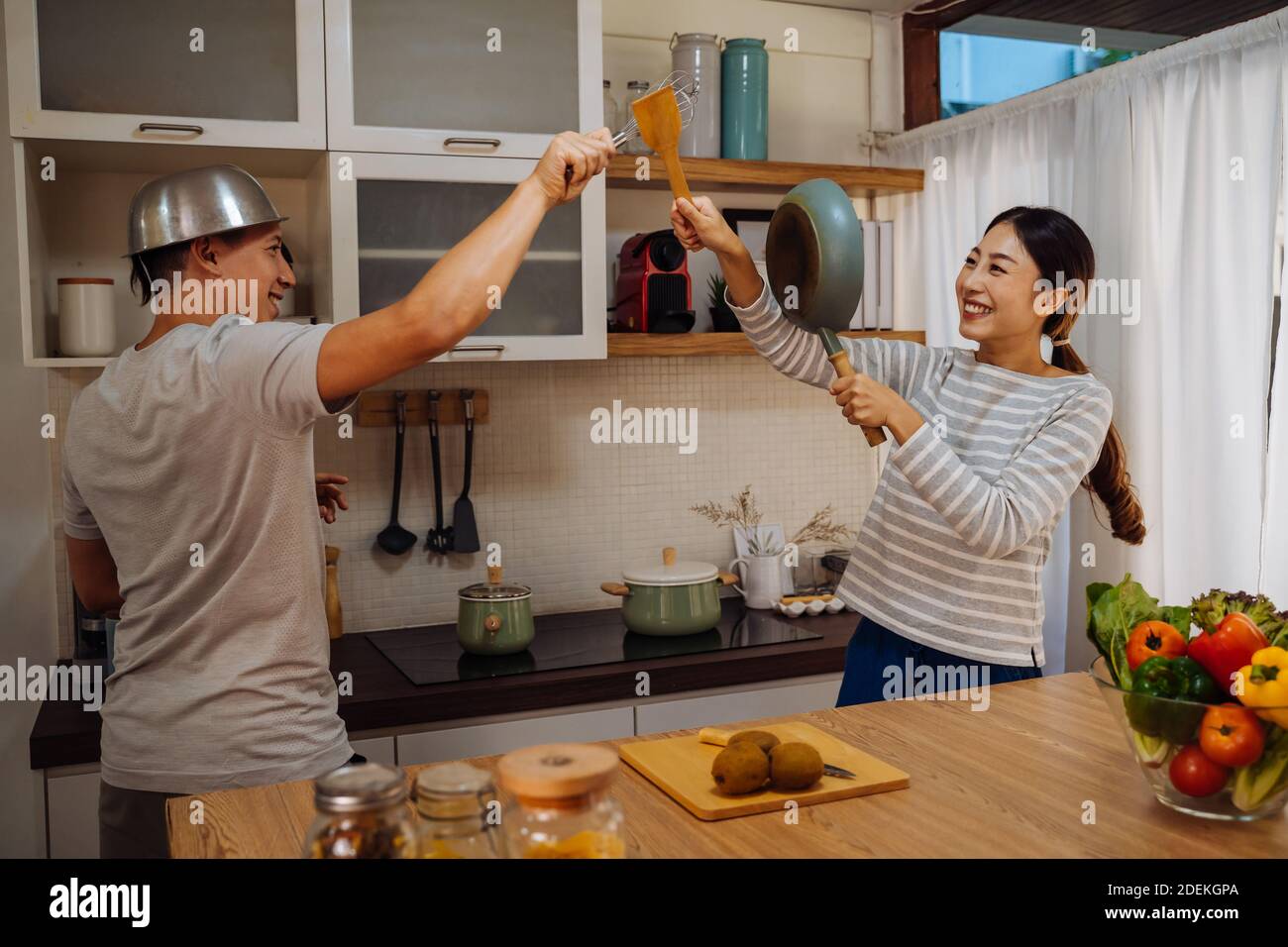 Giovane coppia sposata che fa una lotta informale finding con utensili e padelle in cucina a casa. Marito e moglie divertenti che tengono una cucina divertendosi Foto Stock