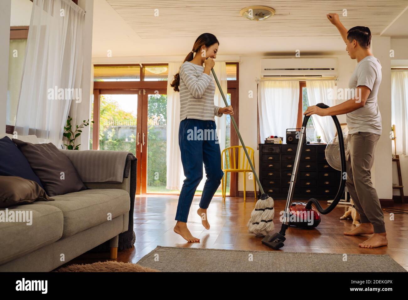 Felice giovane coppia di donne che danzano e cantano durante la pulizia con aspirapolvere e uomo che pulisci il pavimento soggiorno in casa moderna Foto Stock