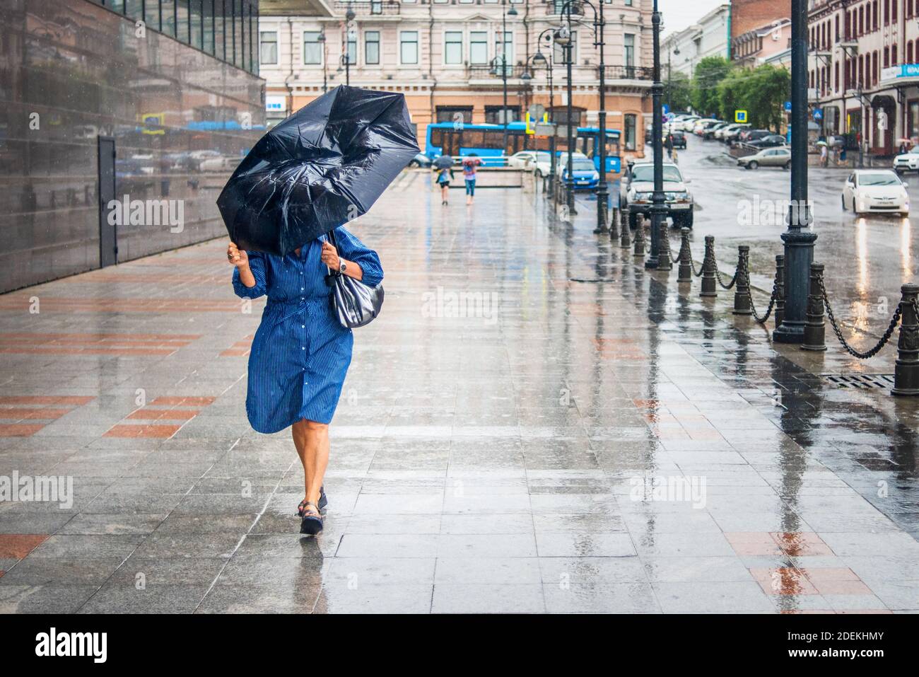 Donna in cattivo tempo piovoso cammina per la strada e cerca di tenere l'ombrello dal vento forte. Paesaggio della città in tempo piovoso. Foto Stock