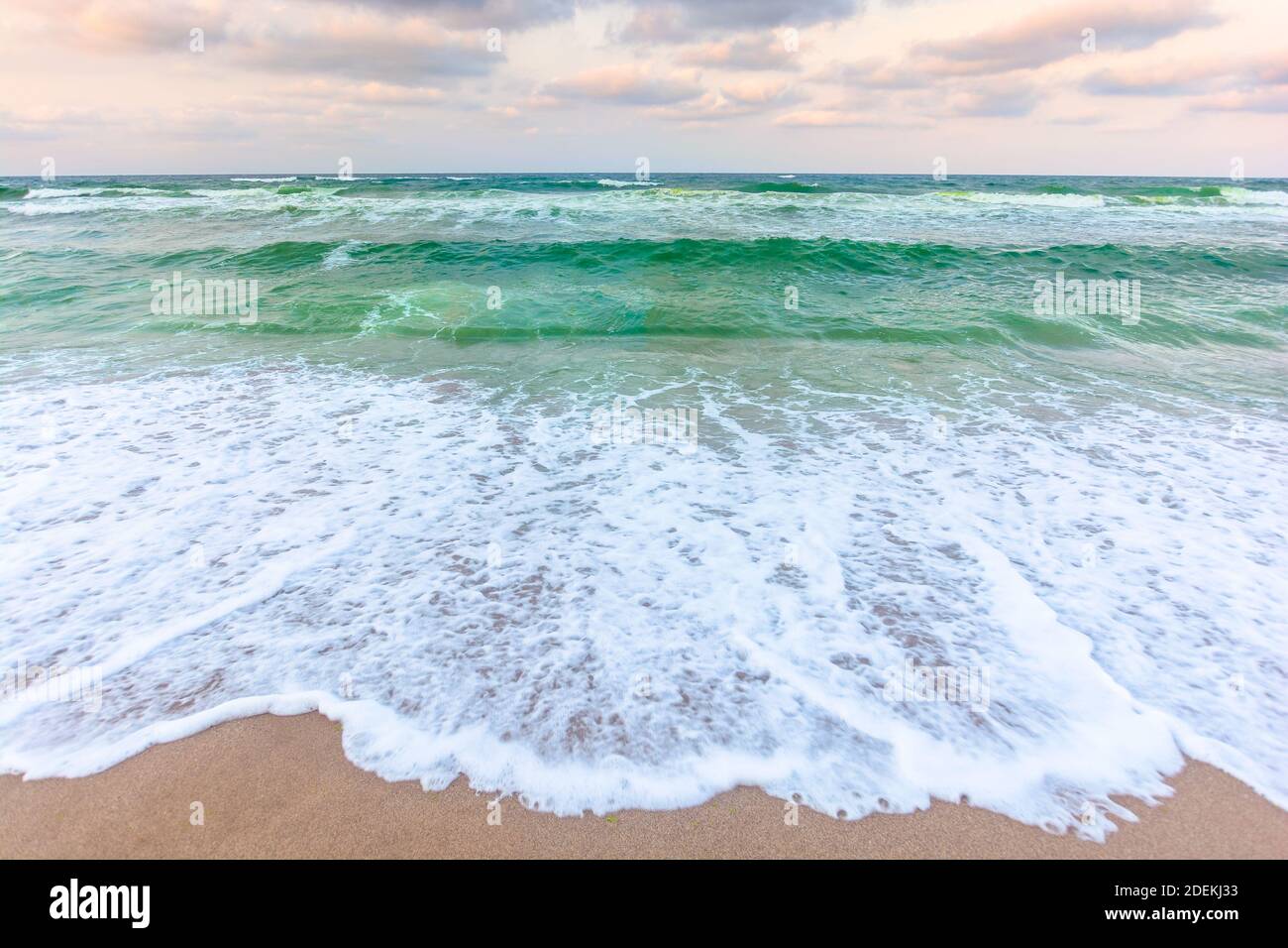 tramonto dorato e nuvoloso sopra le acque verdi del mare. onde che si infrangono sulla spiaggia sabbiosa. orizzonte limpido. tempo ventoso in continuo mutamento Foto Stock