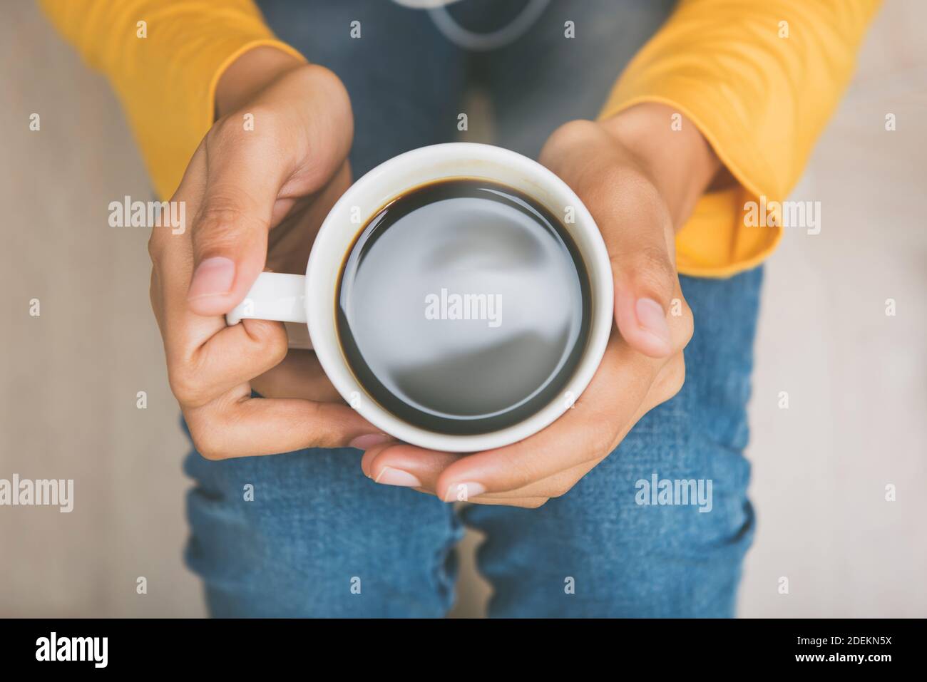 Vista dall'alto delle mani di una giovane donna che tiene una tazza di caldo caffè nero seduto sul pavimento a casa Foto Stock