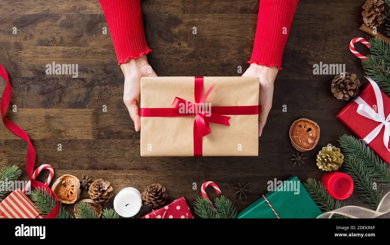 Le mani della donna che mostrano il regalo di Natale sulla tavola di legno circondano con gli articoli e le scatole decoranti, preparati per celebrare la stagione festiva di festa Foto Stock