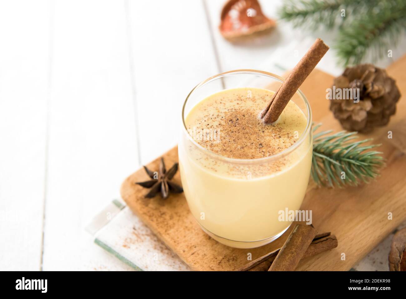 Bevanda casalinga tradizionale di Natale in un bicchiere con noce moscata macinata, cannella e oggetti decorativi su tavola di legno bianco, preparandosi per celebratina Foto Stock