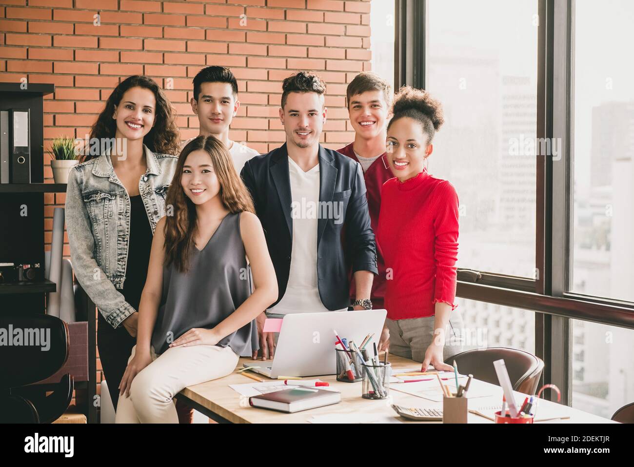 Giovane trendy e variegata squadra millennial sorridente in abiti casual intorno tavolo da ufficio dopo la riunione di affari con muro di mattoni rossi e. finestra skyline della città Foto Stock