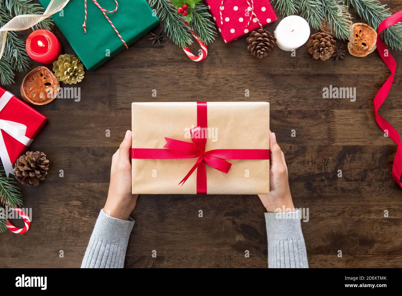 Le mani della donna che mostrano il regalo di Natale sulla tavola di legno circondano con gli articoli e le scatole decoranti, preparati per celebrare la stagione festiva di festa Foto Stock