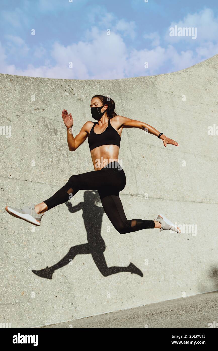 Donna in running e jumping sportswear. Intera lunghezza di donna sana con maschera facciale che si springe all'aperto. Foto Stock