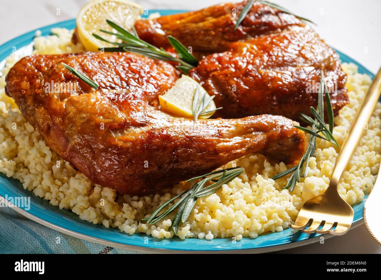 I quarti di gamba di pollo serviti sopra il miglio su un piatto con le posate dorate, i cunei di limone e le sprigs fresche del rosmarino su uno sfondo bianco della pietra di marmo Foto Stock