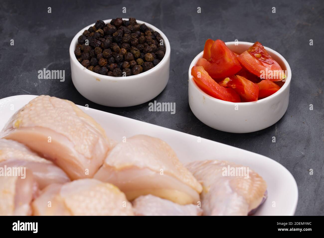 Pollo crudo tenero al curry tagliato con la pelle arrangiata su bianco piatto e guarnito con pepe nero e fettine di pomodoro sfondo di colore grafite Foto Stock
