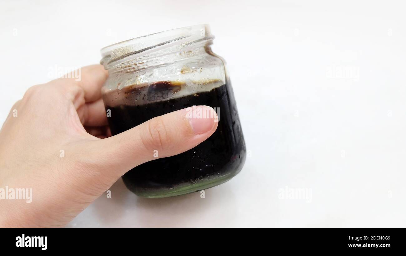 Una mano che tiene un vaso di vetro con gelatina nera all'interno. Senza coperchio. Vista laterale. Foto Stock