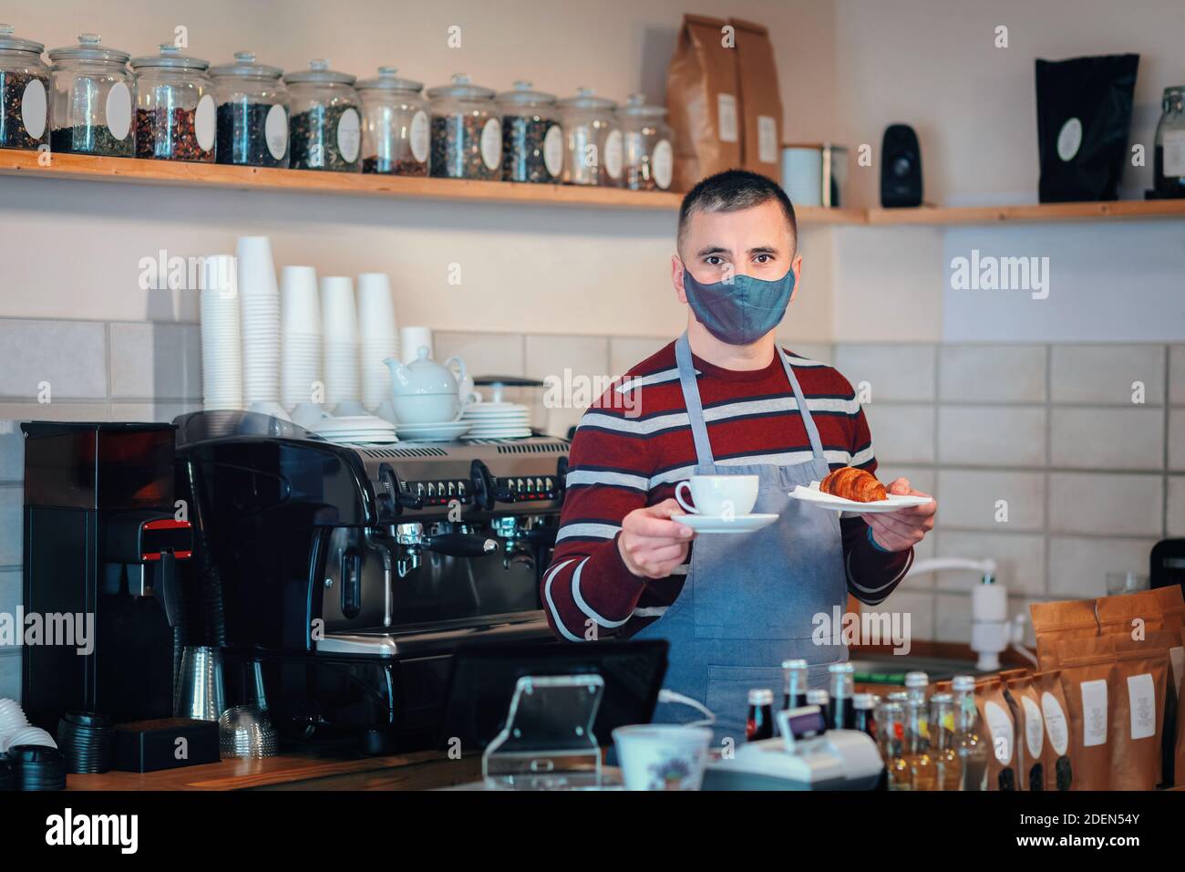 Il proprietario di una piccola azienda indossa una maschera che serve la colazione dietro il bancone Foto Stock