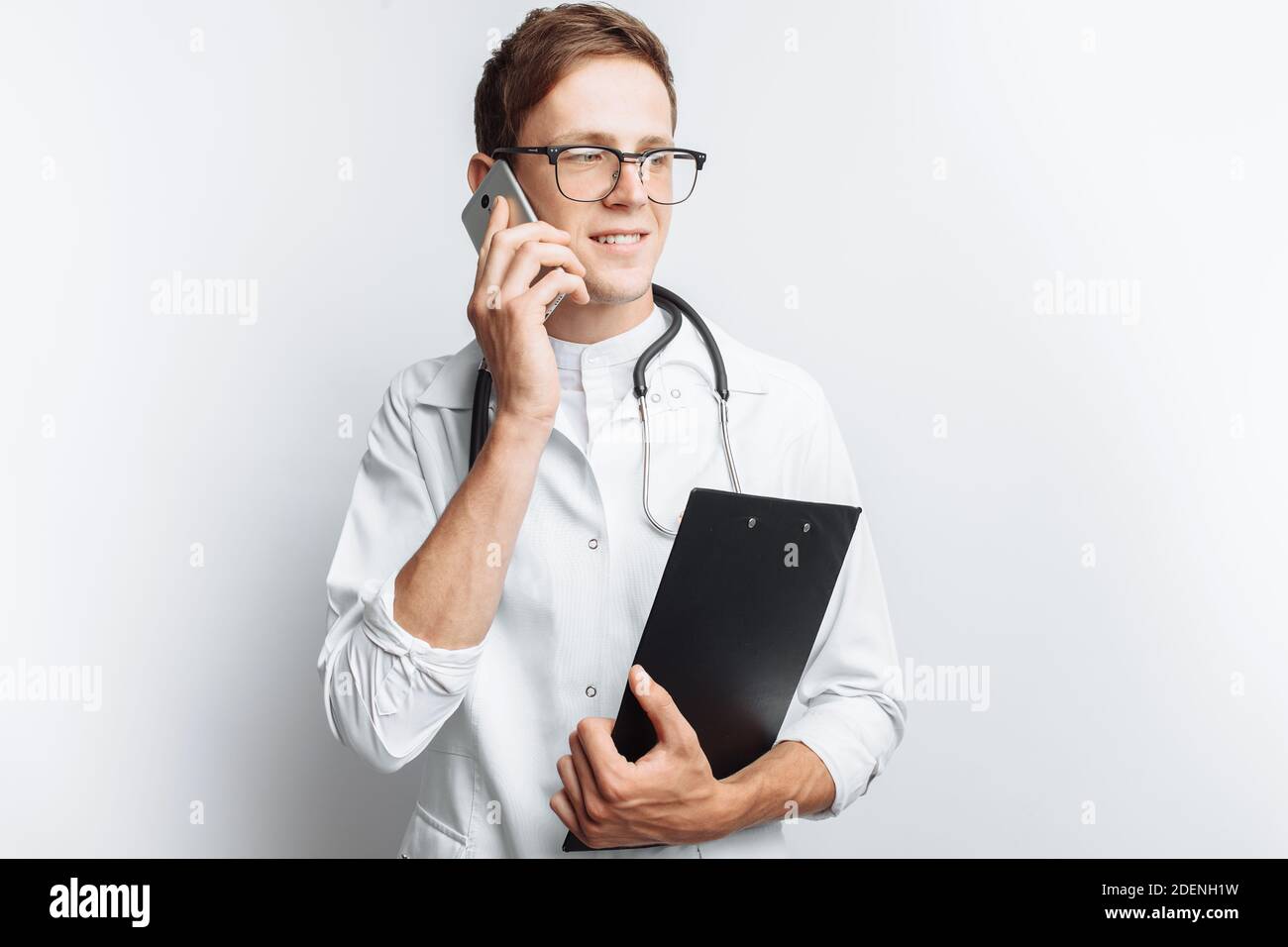 Medico giovane e bello che parla al telefono con i pazienti, studente interno con cartella in mano, sfondo bianco, Foto Stock