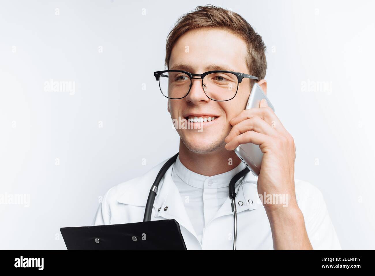 Medico giovane e bello che parla al telefono con i pazienti, studente interno con cartella in mano, sfondo bianco, Foto Stock