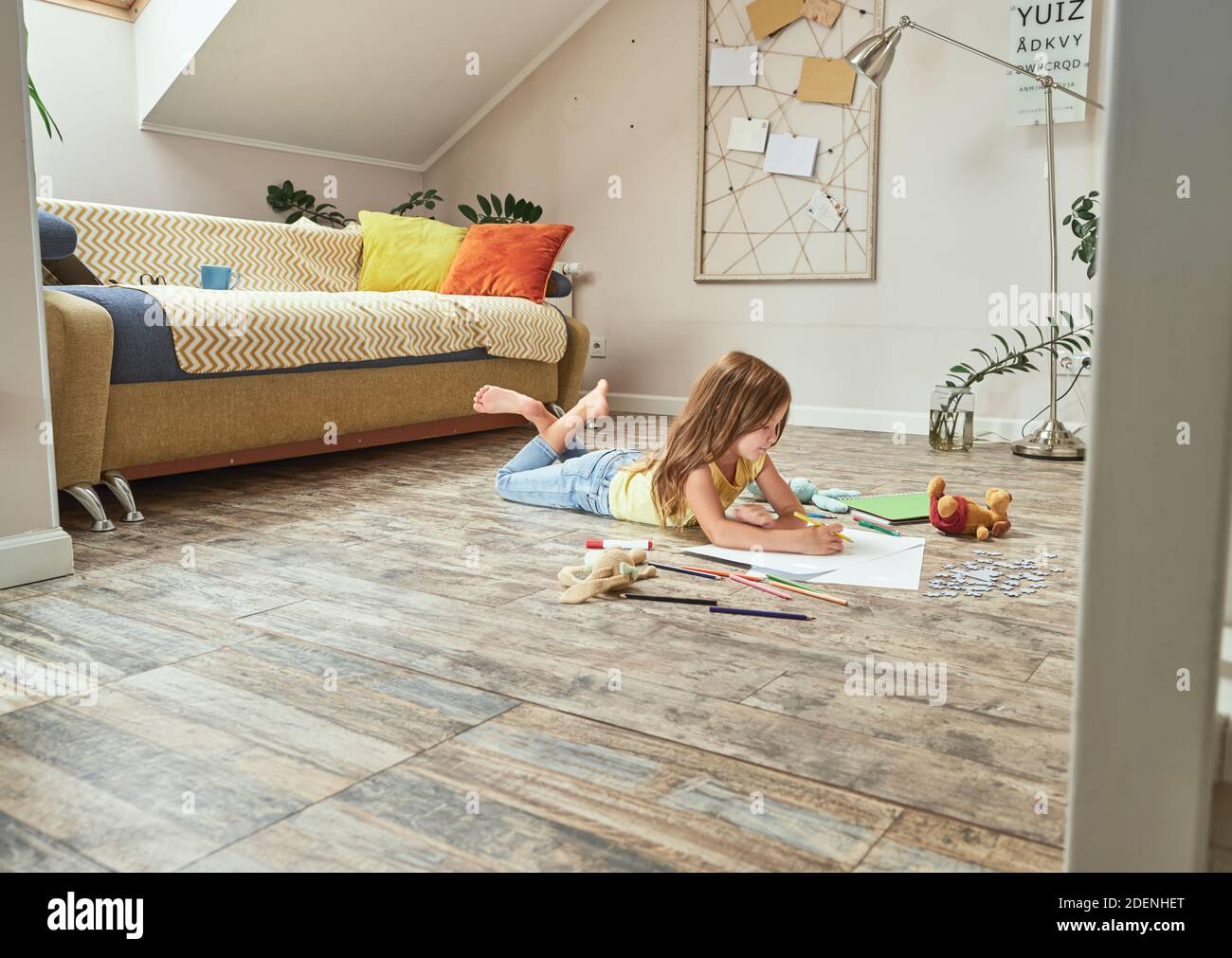 Un bambino creativo che gioca a casa. Piccola ragazza caucasica sdraiata sul pavimento in legno nel soggiorno e disegnata con matite colorate Foto Stock