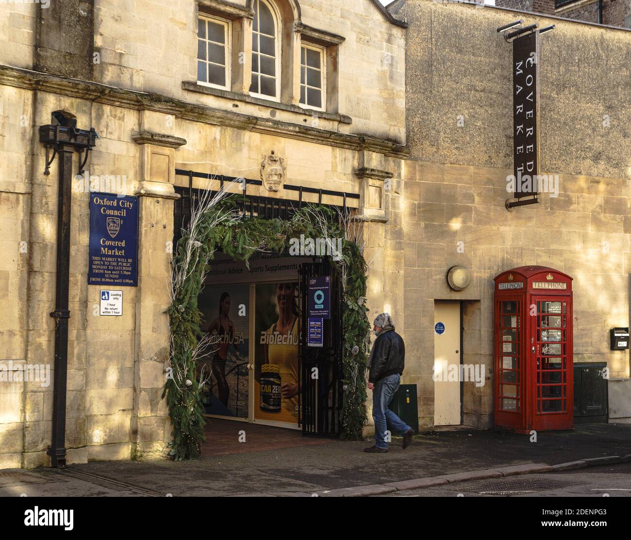 Un uomo di mezza età vestito di jeans e una giacca di pelle, cammina verso l'ingresso decorato al mercato coperto, Oxford, Regno Unito. Natale. Foto Stock