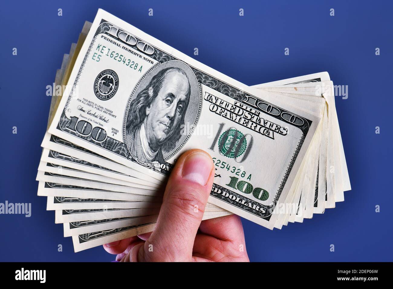 Una mano dell'uomo isolata su uno sfondo blu mostra una mazzo di 1000 dollari in dieci 100 dollari fatture Foto Stock