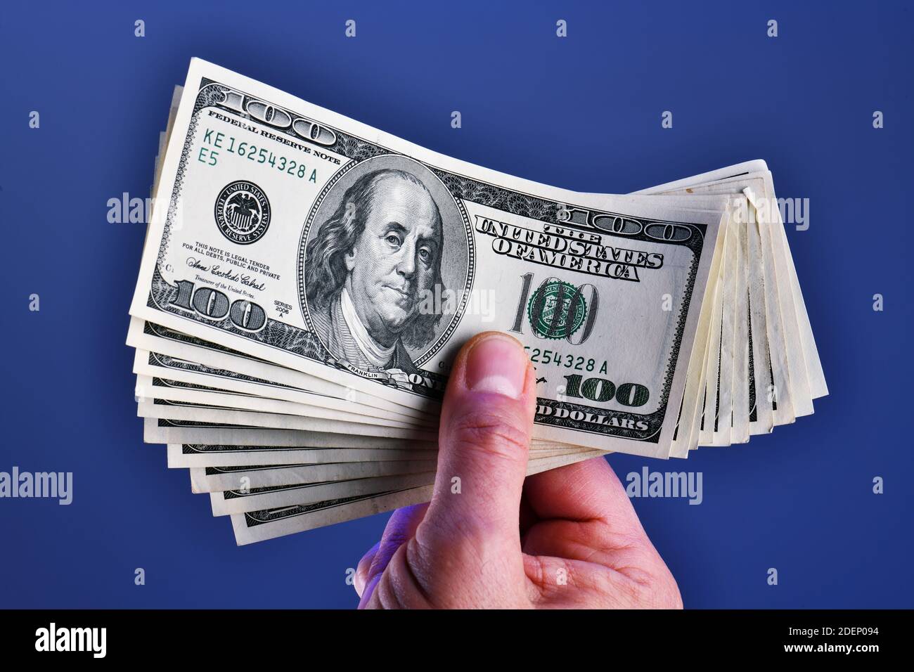 Una mano dell'uomo isolata su uno sfondo blu mostra una mazzo di 1000 dollari in dieci 100 dollari fatture Foto Stock