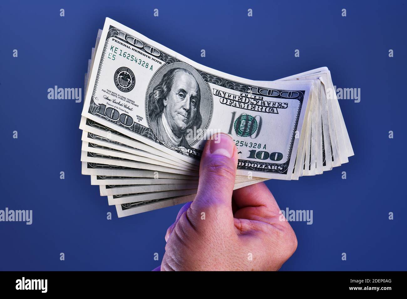 Una mano dell'uomo isolata su uno sfondo blu mostra una mazzo di 1000 dollari in dieci 100 dollari fatture Foto Stock