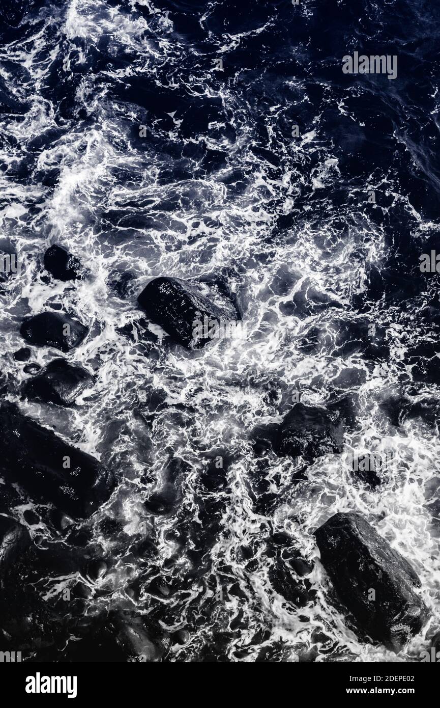 Acqua e rocce (duotone) . Piccole onde si schiantano su una costa rocciosa e creano vortici di schiuma bianca, visti dall'alto Foto Stock