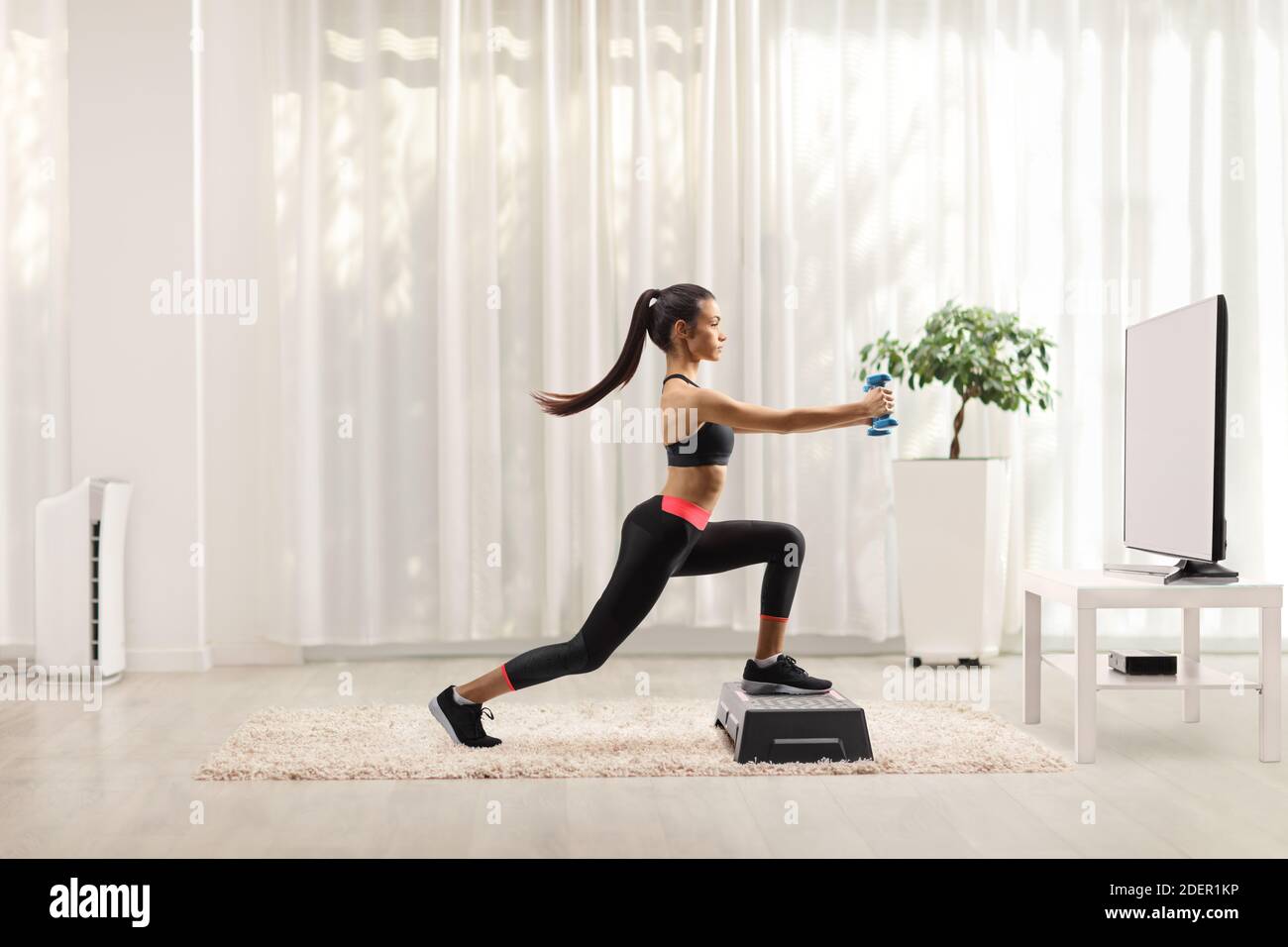 Giovane femmina che esercita passo aerobico con piccoli pesi davanti di un televisore a casa Foto Stock