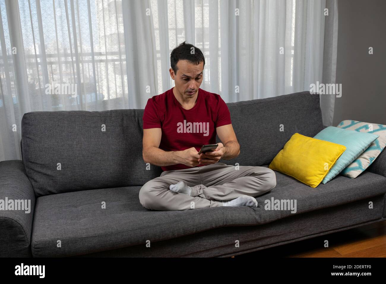 Uomo maturo (44 anni) seduto con le gambe incrociate sul divano e molto sorpreso di vedere il suo smartphone. Foto Stock