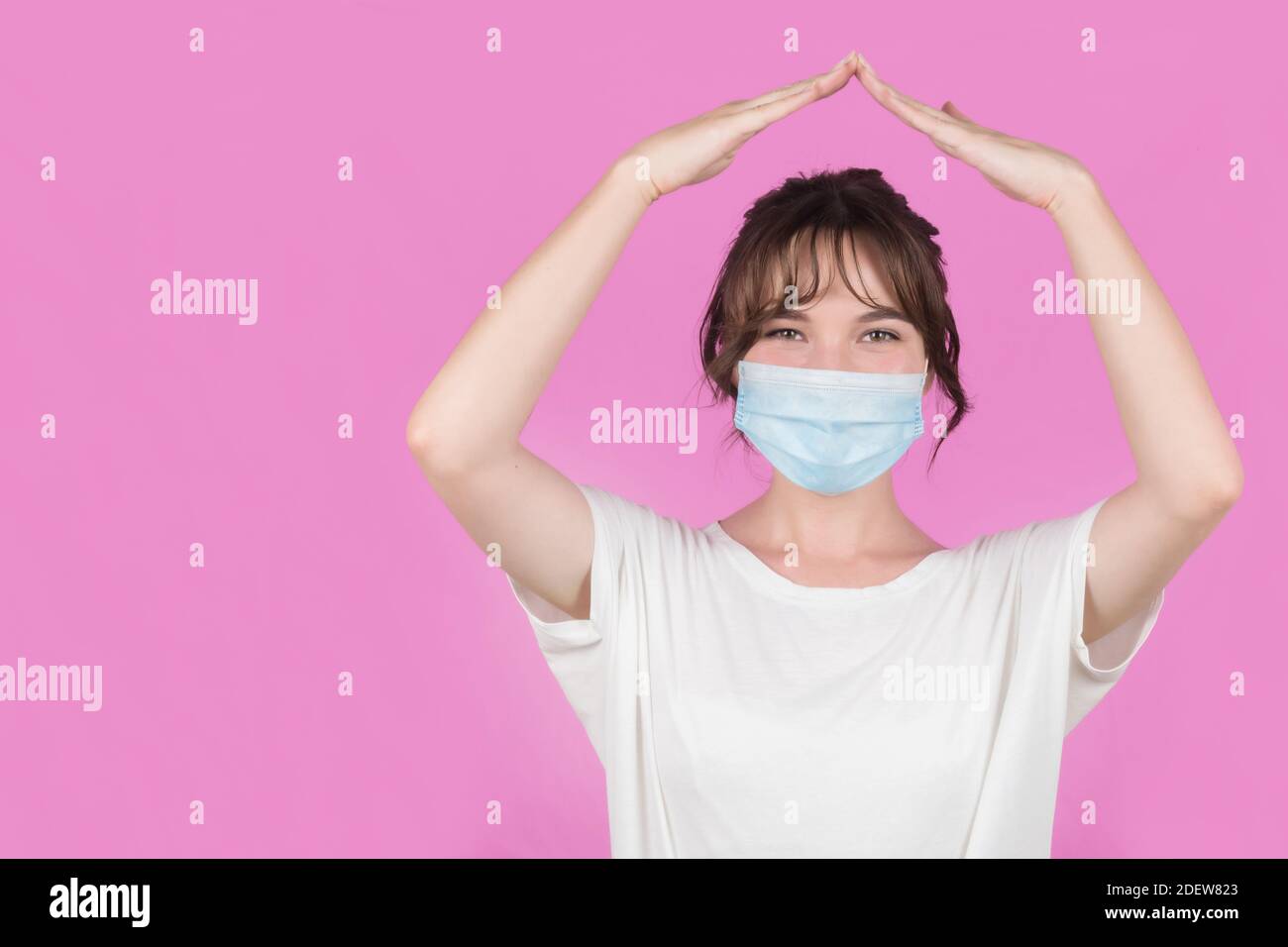 Ritratto di una giovane donna in una maschera medica che fa un gesto domestico. Rimani a casa. Sfondo rosa. Foto di alta qualità Foto Stock