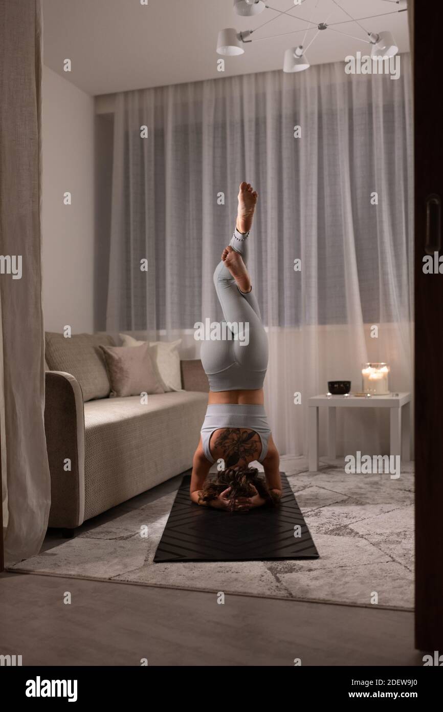 Vista posteriore di una donna anonima che fa supportato Headstand con Eagle Gambe a casa Foto Stock