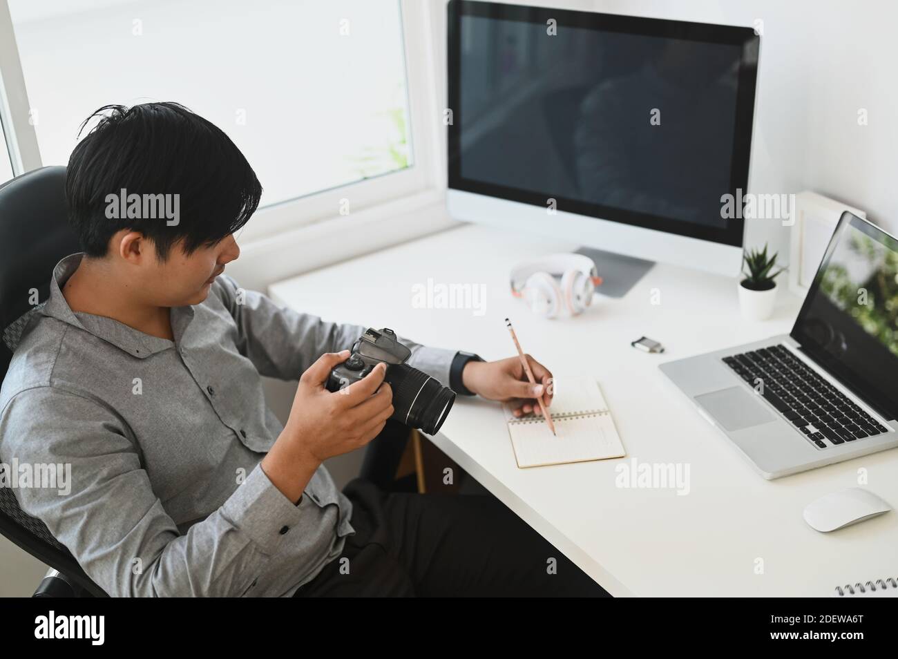 Un giovane fotografo che riesamina le sue foto scattate con la sua fotocamera mentre si siede alla sua scrivania all'interno dello studio. Foto Stock