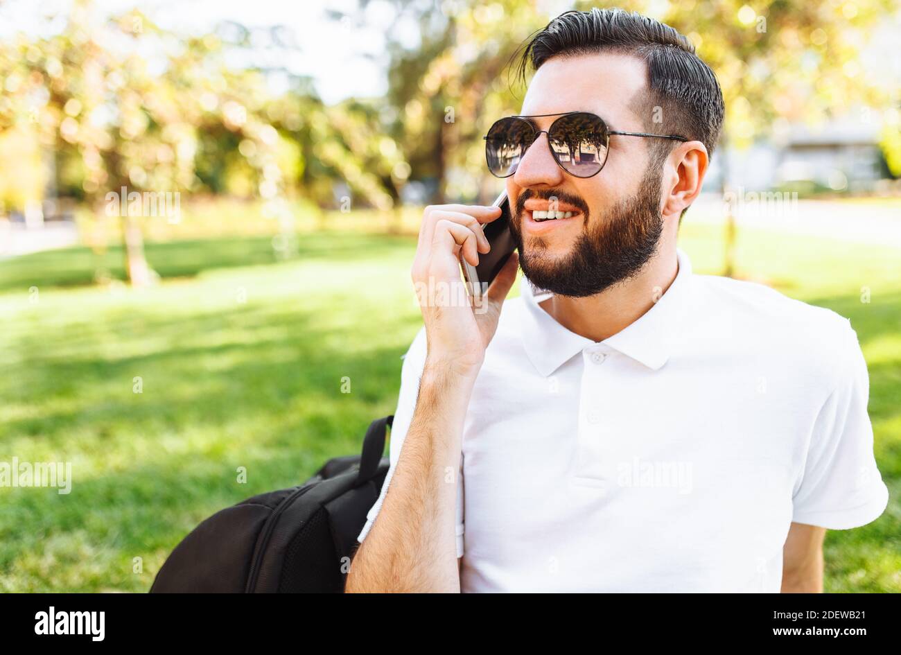 Hipster business ed elegante in una camicia bianca e occhiali da sole, seduto sul prato nel Parco e parlando al telefono Foto Stock