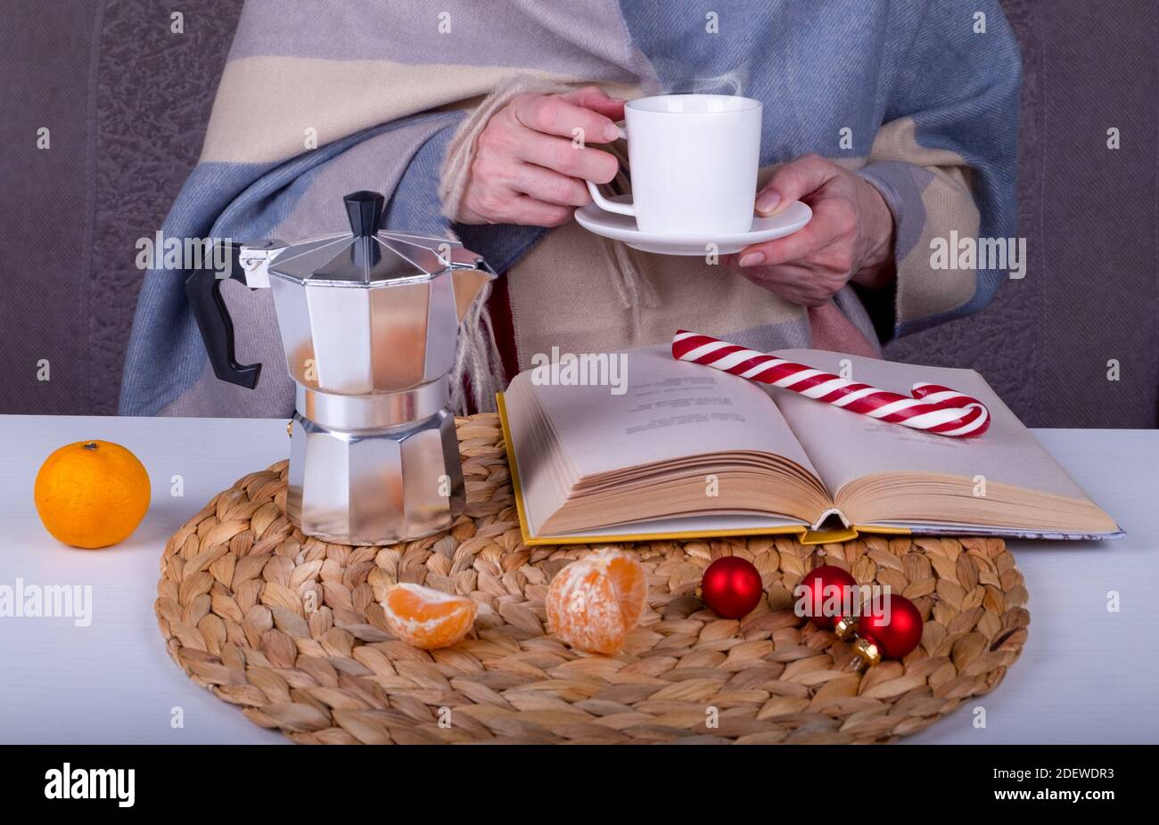 Primo piano con un libro, una macchina per il caffè geyser, decorazioni natalizie sul tavolo. Una donna tiene una tazza bianca con una bevanda calda del caffè. Foc. Selettivo Foto Stock