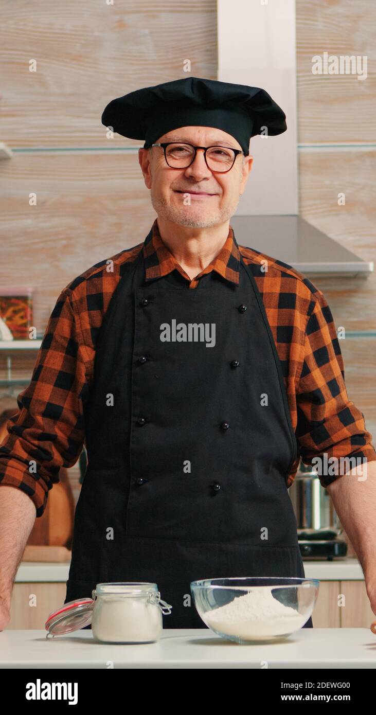 Ritratto dello chef che indossa bonete guardando la fotocamera e sorridendo. Panettiere anziani in cucina uniforme preparare ingredienti di pasticceria su tavola di legno pronti a cucinare pane, torte e pasta gustosi fatti in casa Foto Stock