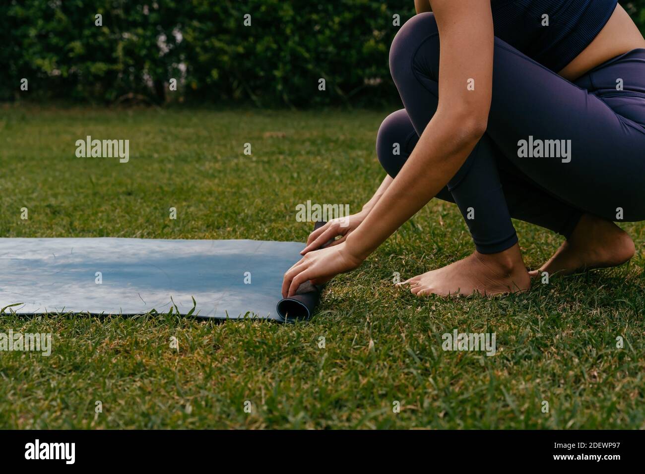 Praticante di yoga femminile a piedi nudi che srotola il tappetino sull'erba mentre si prepara per lezioni di yoga in cortile Foto Stock