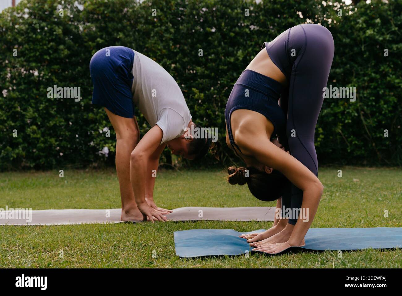 Vista laterale dell'uomo e della donna che fanno la curva in avanti in piedi sui tappetini mentre si pratica yoga in cortile il giorno d'estate Foto Stock