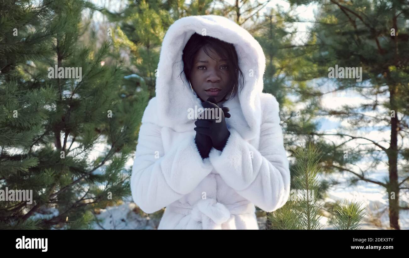 triste donna dalla pelle scura tremola con freddo e riscalda le mani contro pinete verdi in inverno freddo primo piano Foto Stock