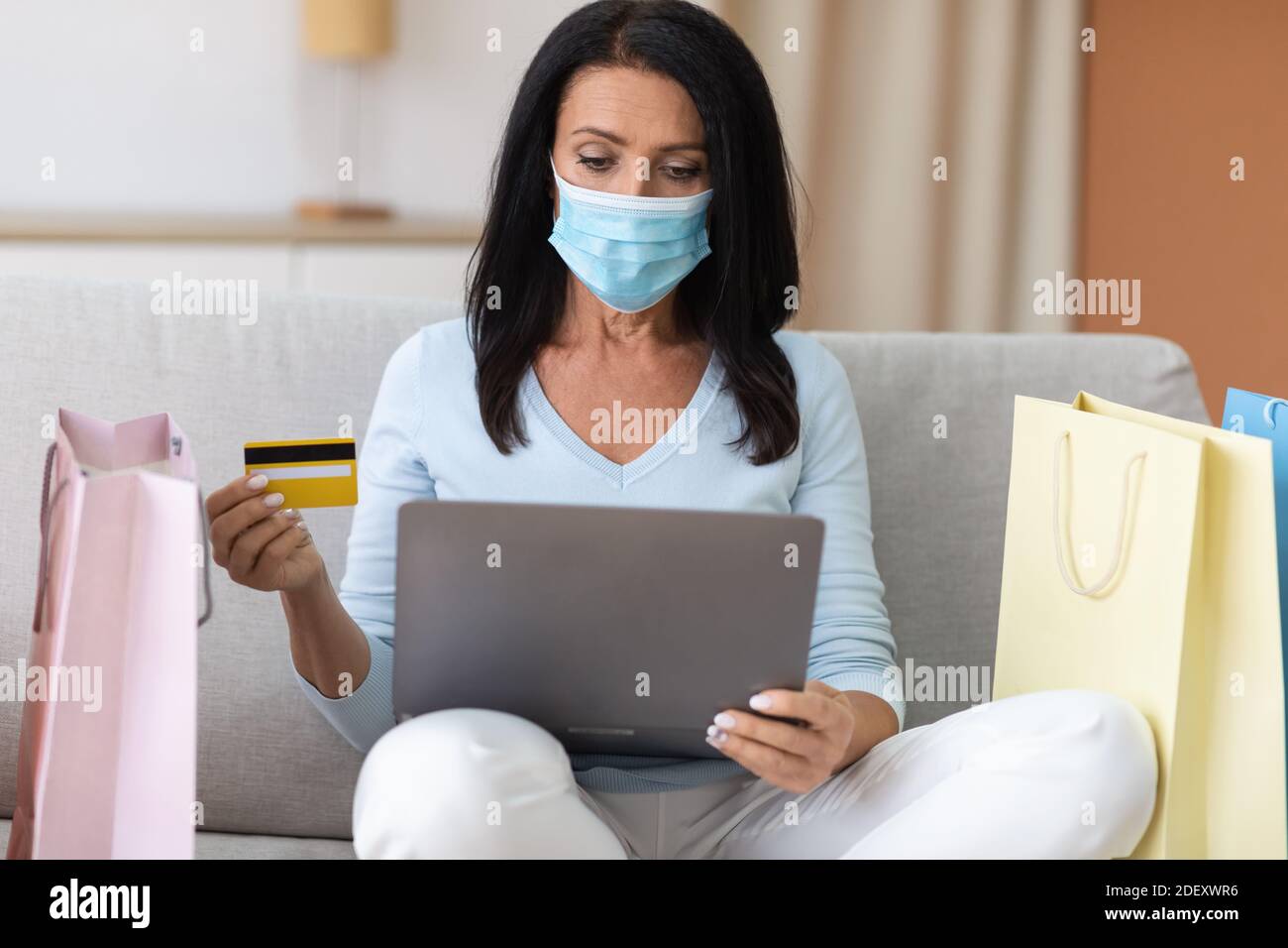 Signora matura in maschera medica utilizzando il computer portatile e la carta di credito Foto Stock