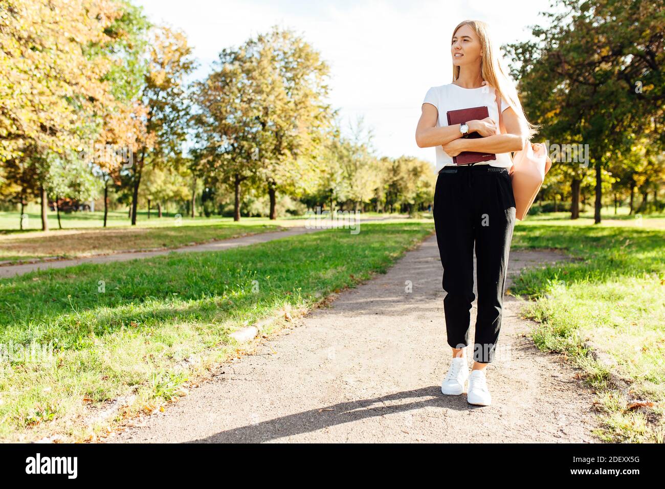 Bella studentessa che cammina nel parco tenendo un libro dentro un buon umore va a casa dopo la scuola Foto Stock