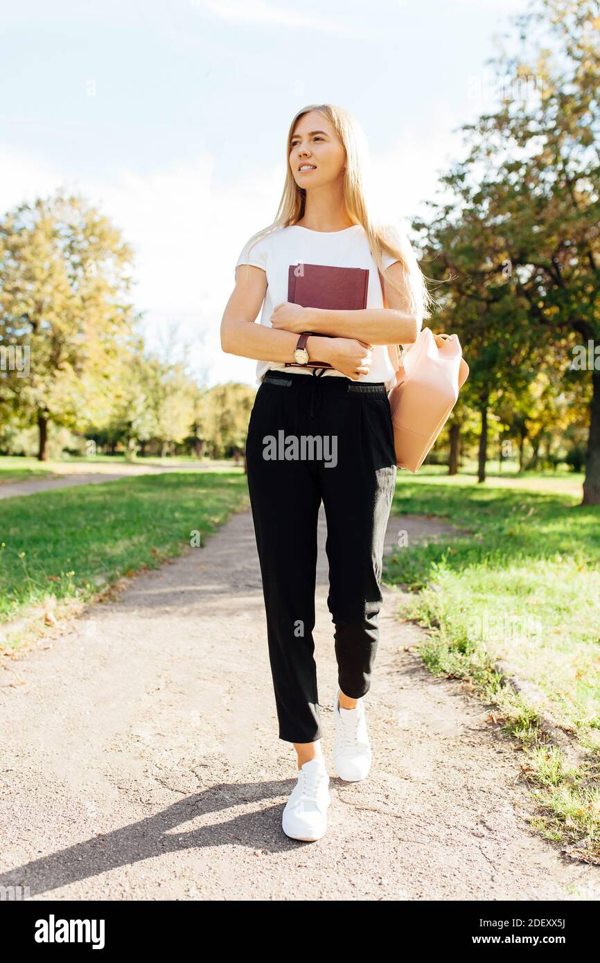 Bella studentessa che cammina nel parco tenendo un libro dentro un buon umore va a casa dopo la scuola Foto Stock